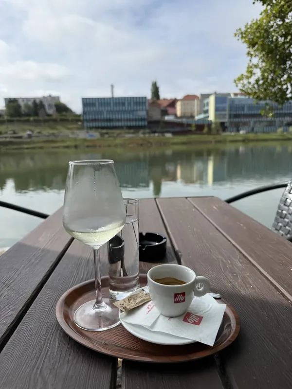 Kozarec vina in skodelica kave na leseni mizi terase, v ozadju mirna Drava in sodobna arhitektura Maribora
