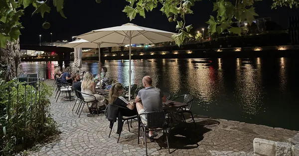 Gostje na večerni terasi Vinoteke Maribor ob reki Dravi, v ozadju osvetljen most