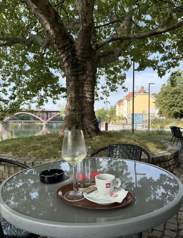Kozarec belega vina in skodelica kave illy na mizi pod platano, v ozadju most čez Dravo in mestne stavbe